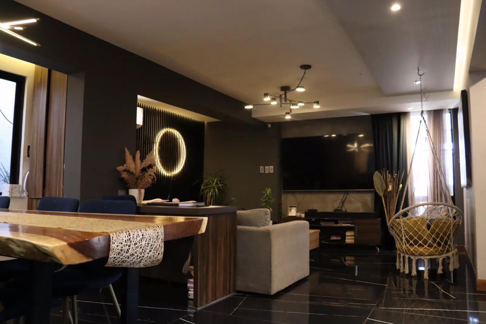 Comedor de madera natural con un espejo circular iluminado al fondo y la sala detrás.