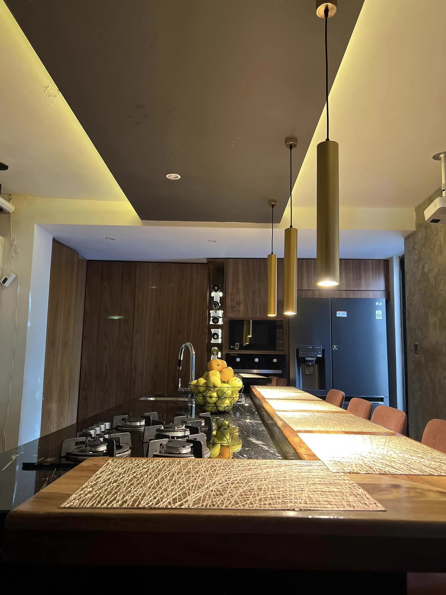 Cocina de Casa 1009, con isla de granito negro, barra de madera y lámparas colgantes doradas bajo un plafón con luz indirecta.