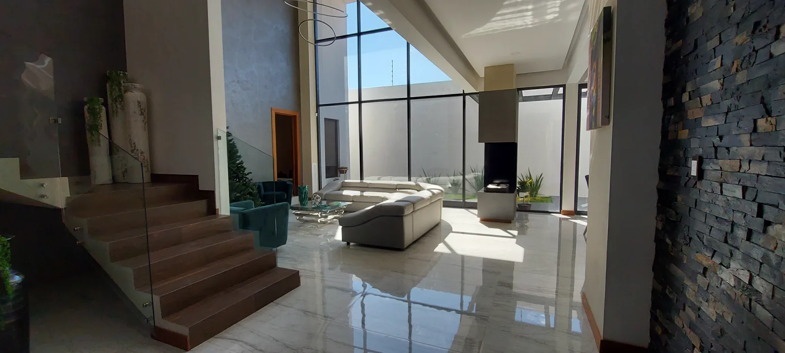 Sala de doble altura con escalera de madera y cristal, muro de piedra laja y chimenea.