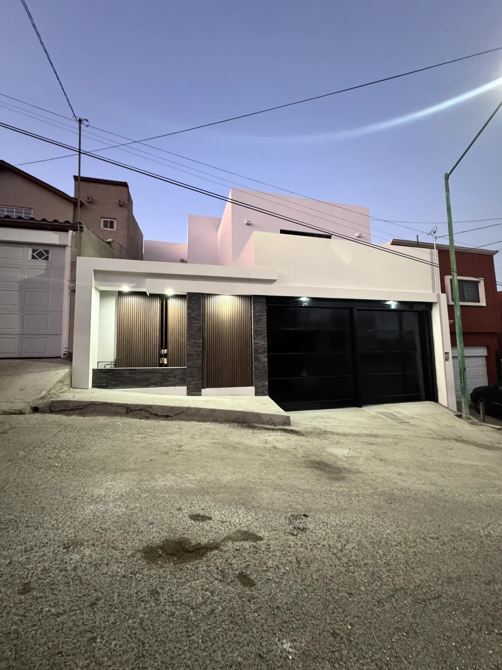 Fachada terminada al atardecer, con la iluminación del acceso encendida y el portón de cristal negro.