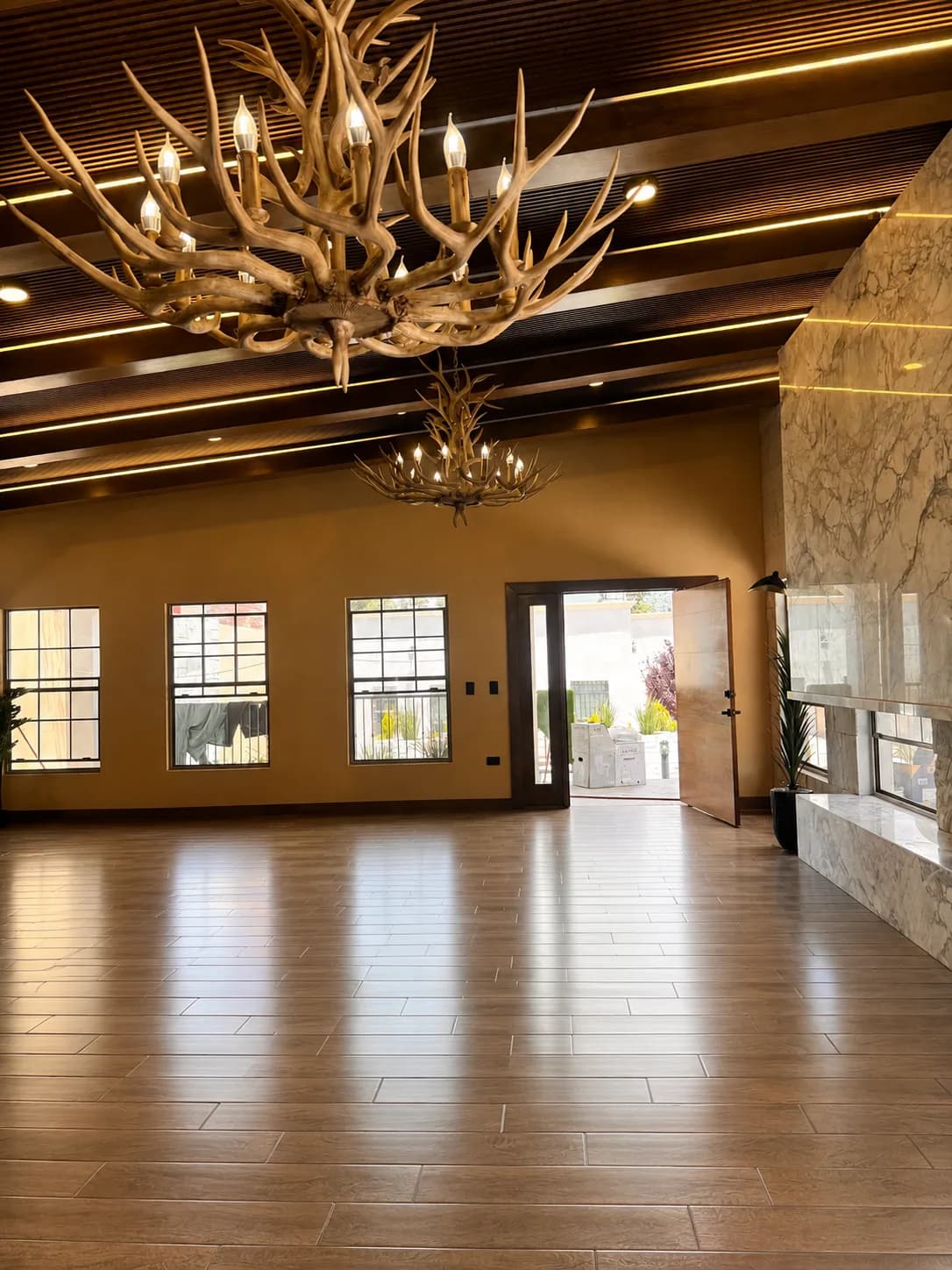Salón con candiles de astas, ventanas corridas y la puerta abierta hacia el exterior.