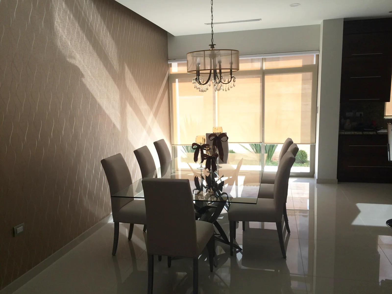 Comedor con mesa de cristal, sillas tapizadas, candil de pantalla y muro de papel tapiz con relieve.