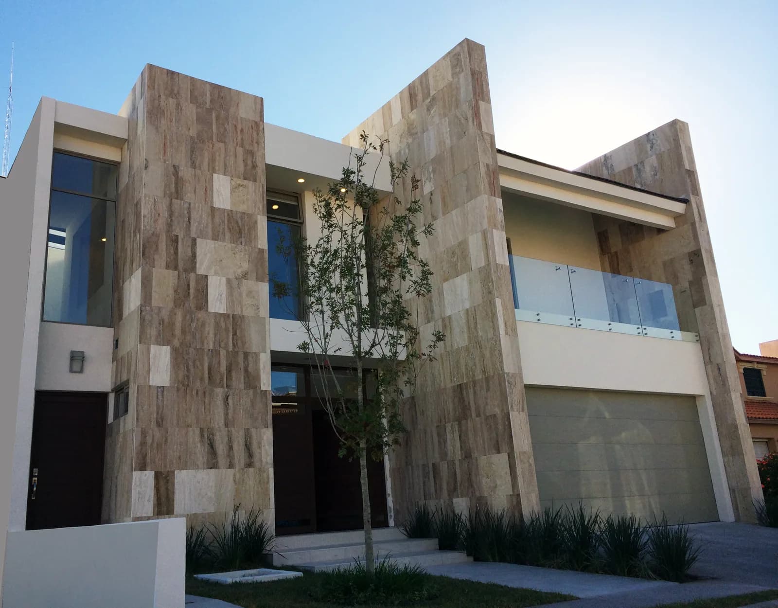 Fachada de Casa Haciendas con placas de travertino, balcón con barandal de cristal y jardín de agaves.