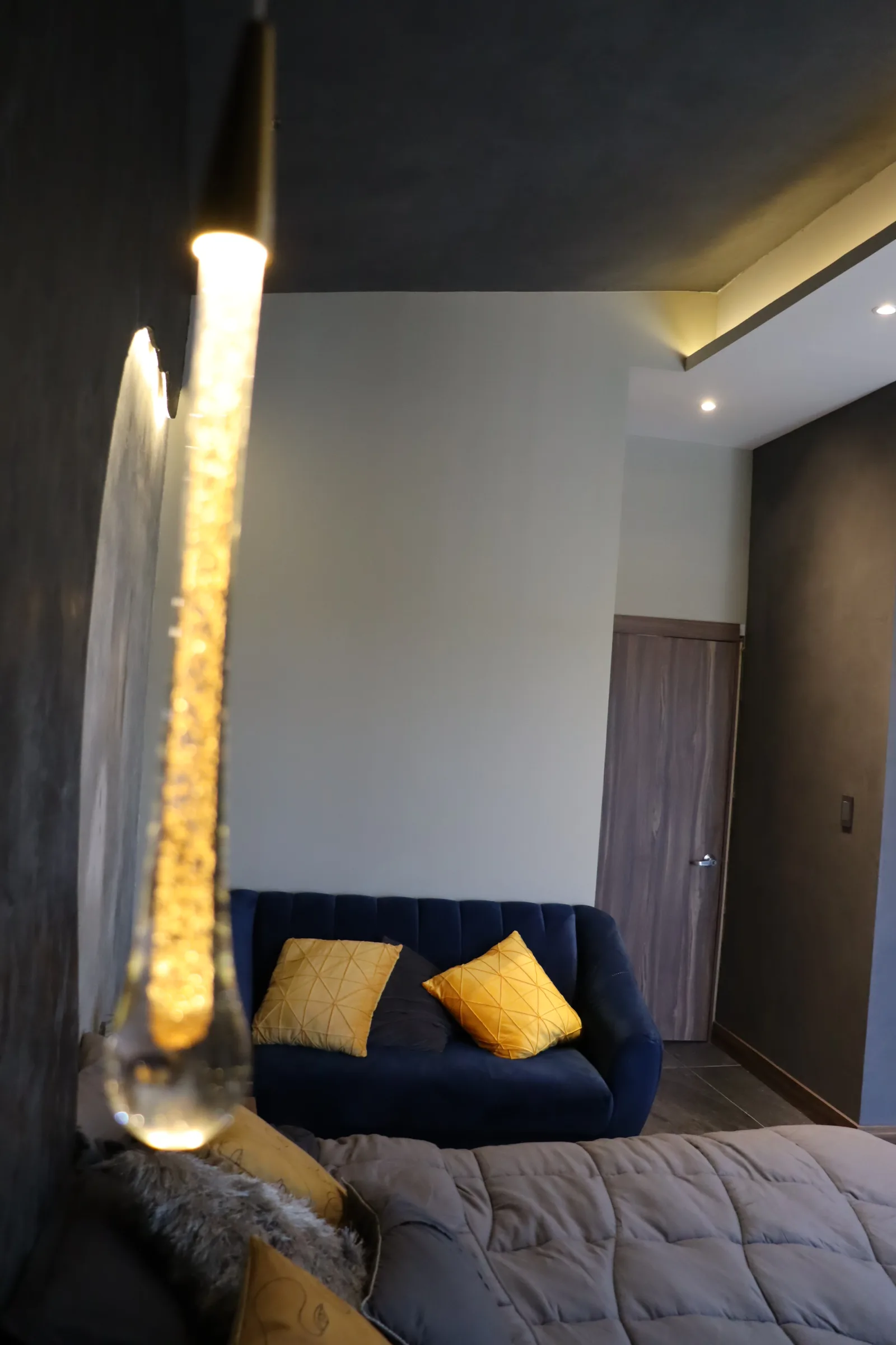 Lámpara de gota en primer plano, con el sillón azul y la recámara al fondo.