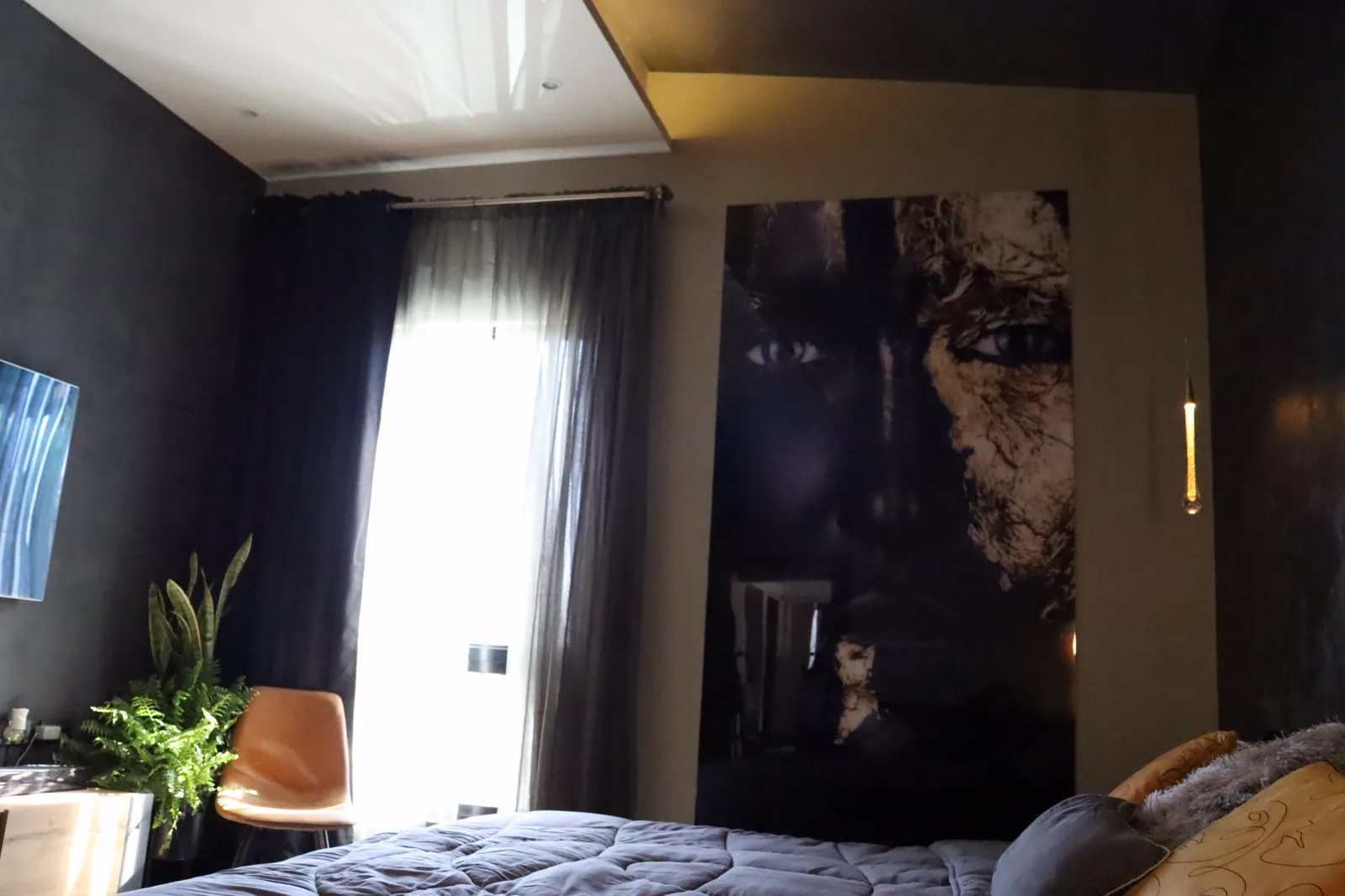 Ventanal con cortinas oscuras, silla de piel y plantas junto a la cama.