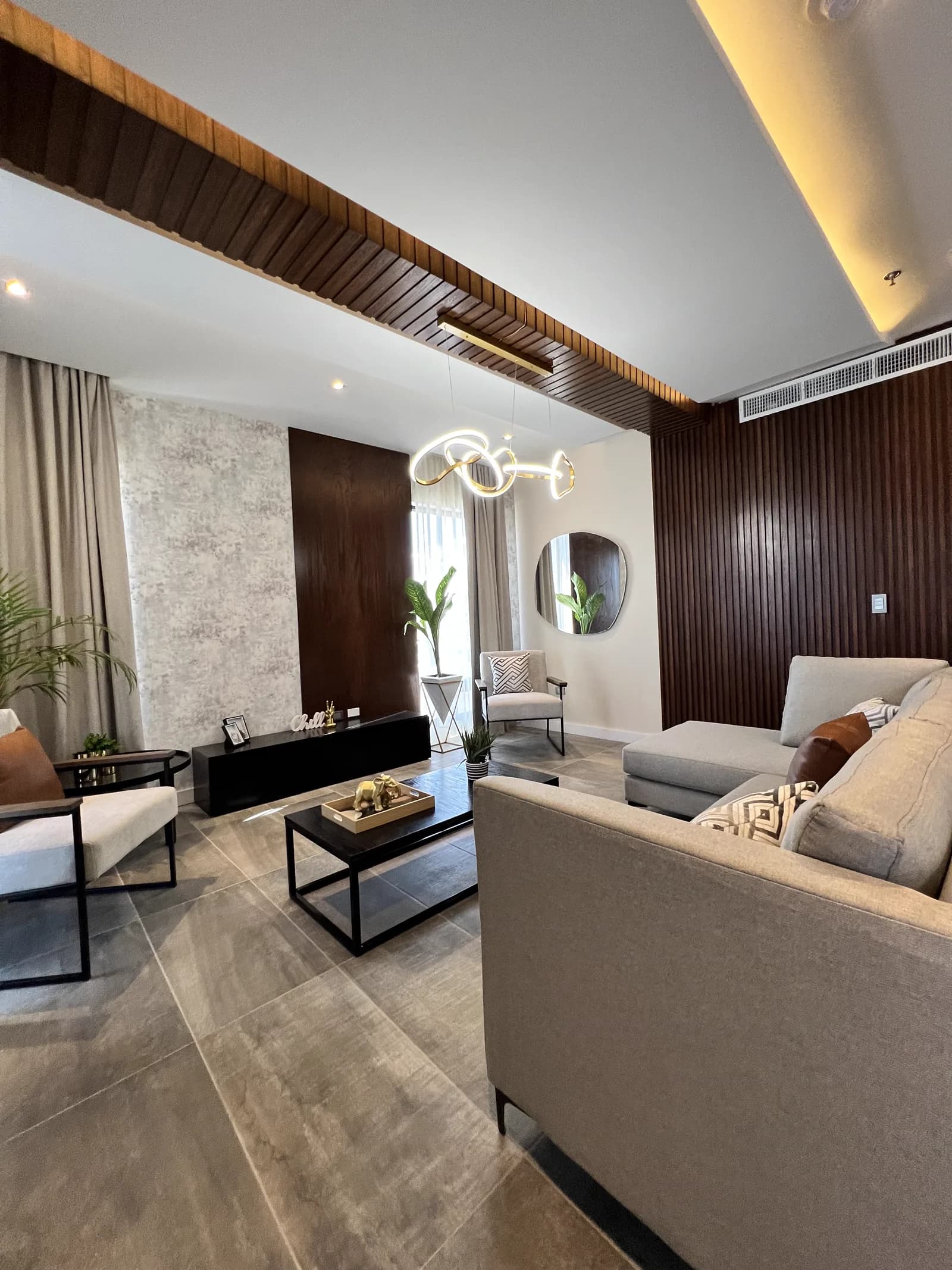 Sala de Departamento All Loft 401, con sillón gris, muro de listones de madera oscura y lámpara de trazo libre.