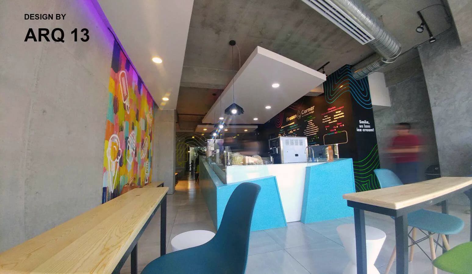 Vista a lo largo del local, con la barra turquesa, mesas de madera clara y sillas de colores.