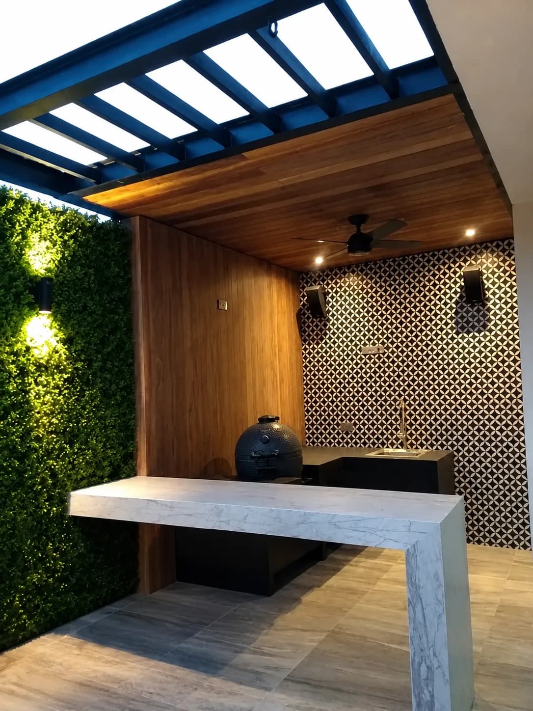 Área de asador de Patio OSR, con barra de piedra blanca, muro de madera, mosaico geométrico y techumbre de cristal.