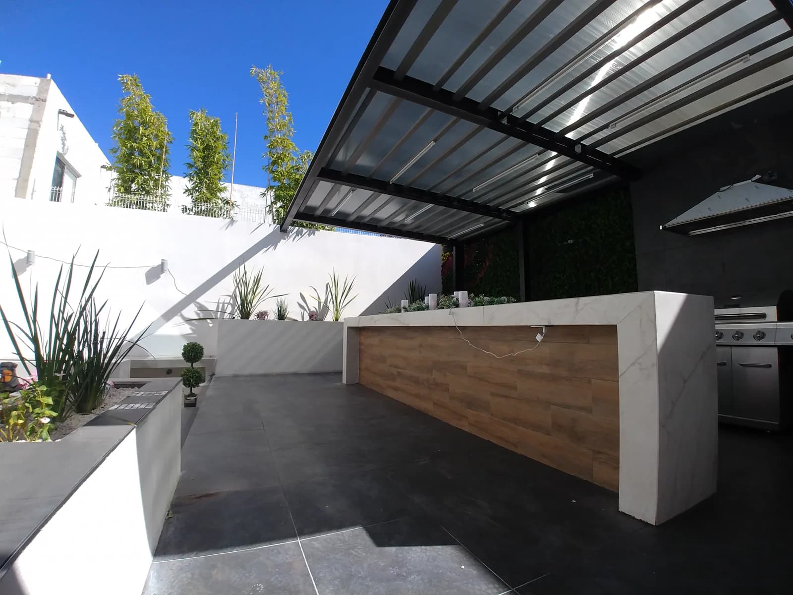 Barra de Patio Rinconadas, de porcelanato tipo mármol y madera, bajo la pérgola de vigas metálicas negras.