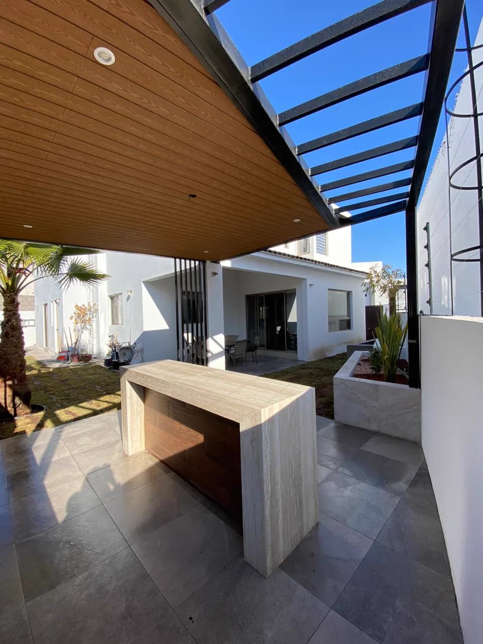 Barra de travertino bajo la pérgola, con la casa al fondo.