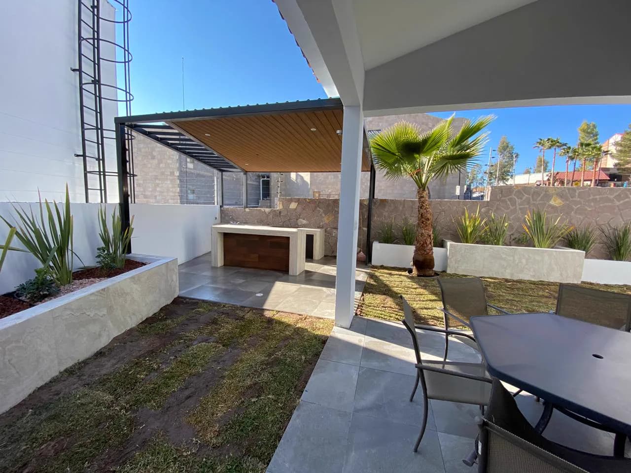 Terraza de Patio San Charbel, con pérgola negra, plafón de madera, barra de travertino y comedor exterior.
