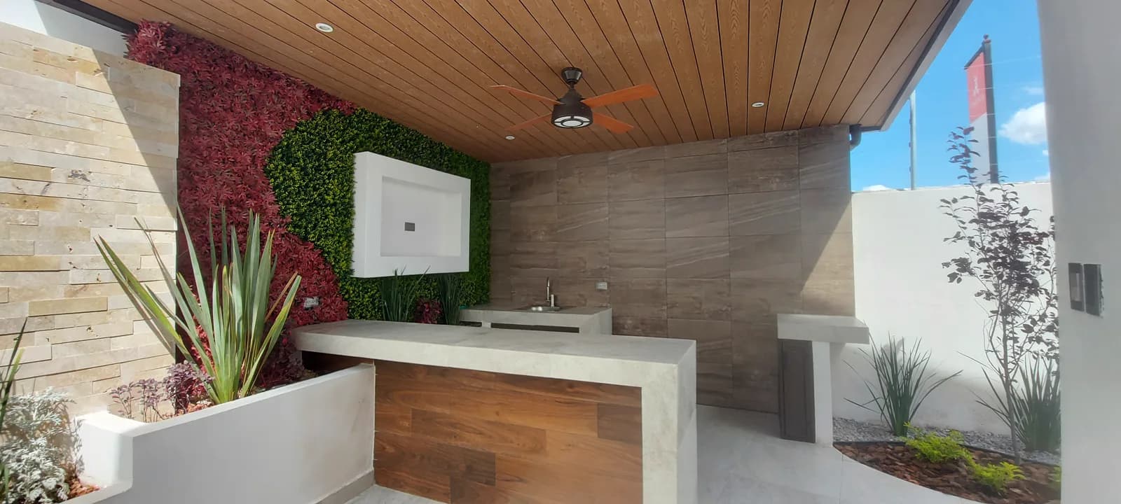 Terraza techada con plafón de madera, barra de cubierta clara y muro verde con follaje rojo.